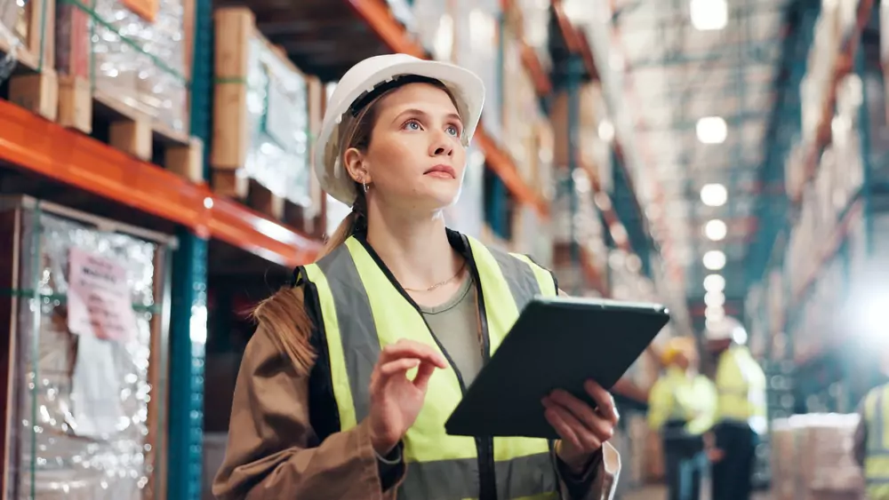 Female warehouse worker in safety vest and hard hat using tablet for inventory management