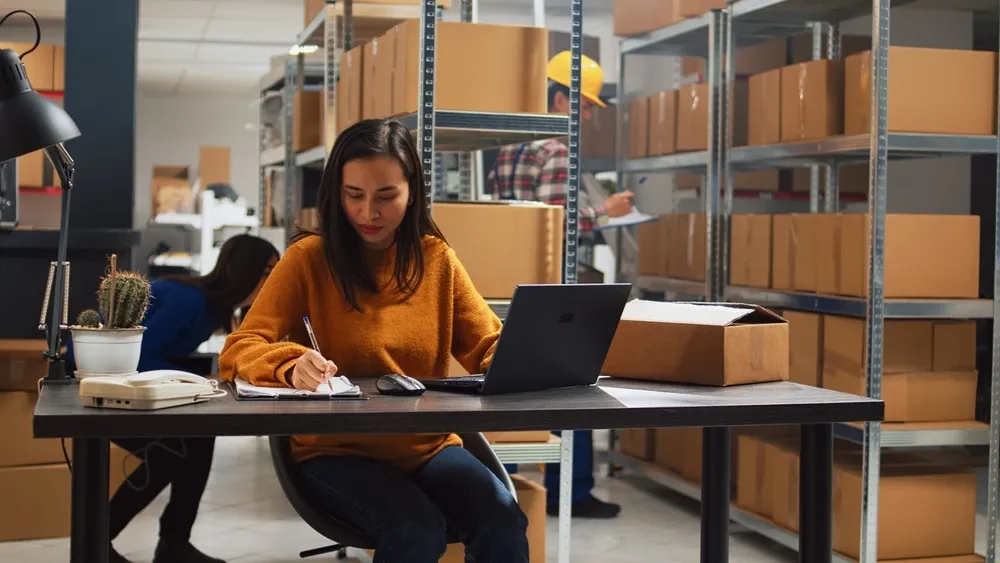 Woman working on laptop in warehouse with automated storage shelving systems and inventory boxes