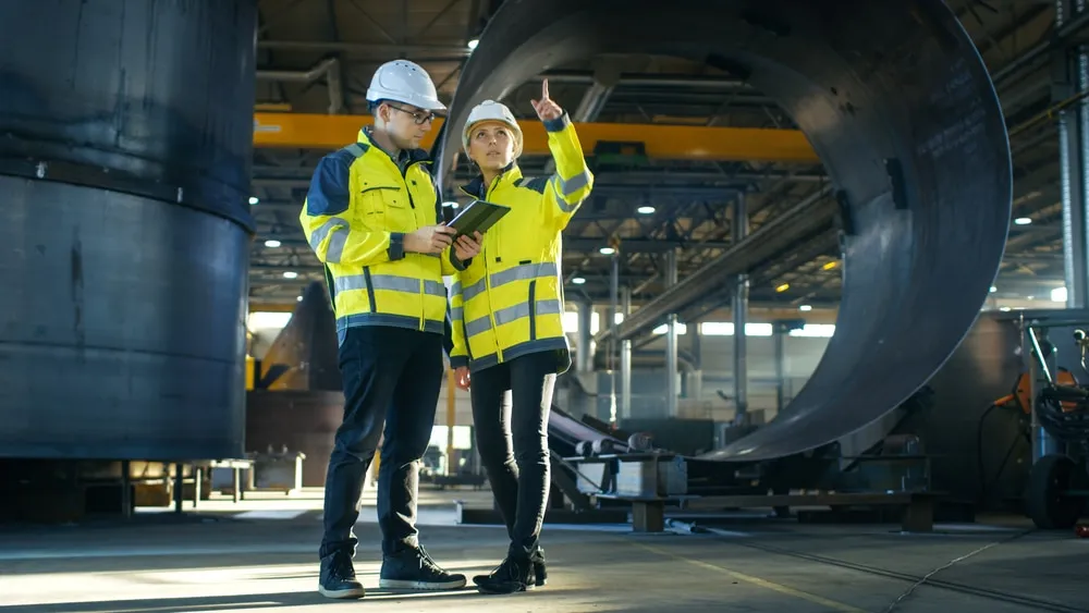 Two manufacturing engineers in safety gear reviewing production schedules on tablet in industrial facility